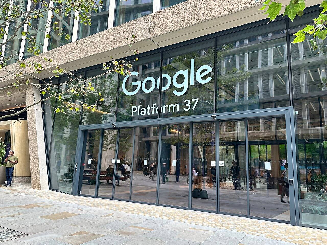 Rótulo iluminado «Google Platform 37» sobre la fachada acristalada de la entrada principal del campus de Google en King's Cross, Londres, con transeúntes junto a las puertas correderas