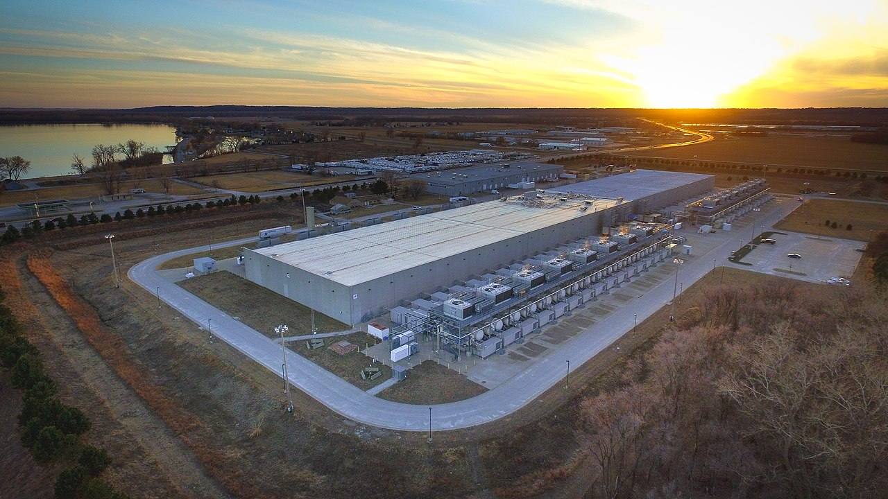 Vista aérea al atardecer del centro de datos de Google en Council Bluffs (Iowa): nave industrial alargada con una larga fila de unidades de refrigeración junto a la fachada, rodeada de campo abierto y con un lago al fondo