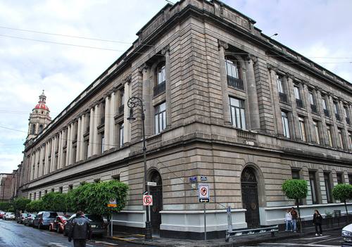 Esquina del edificio histórico de la Secretaría de Educación Pública (SEP) en el Centro Histórico de la Ciudad de México, con la torre de un antiguo convento al fondo y peatones en la acera.
