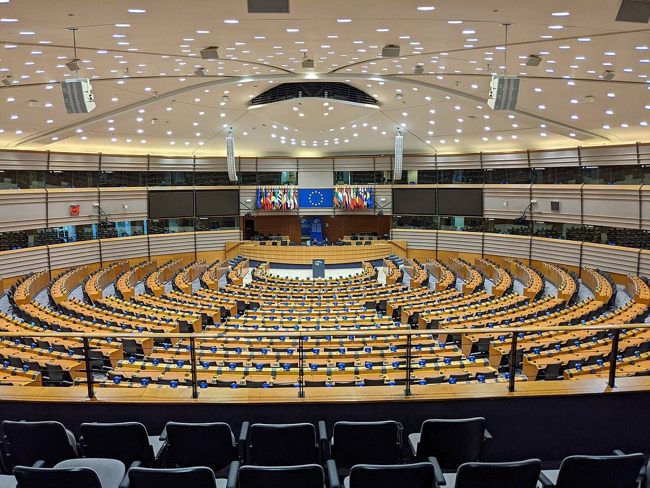 Hemiciclo vacío del Parlamento Europeo en Bruselas, filas de escaños azules numerados y el emblema de estrellas de la UE en el atril del fondo