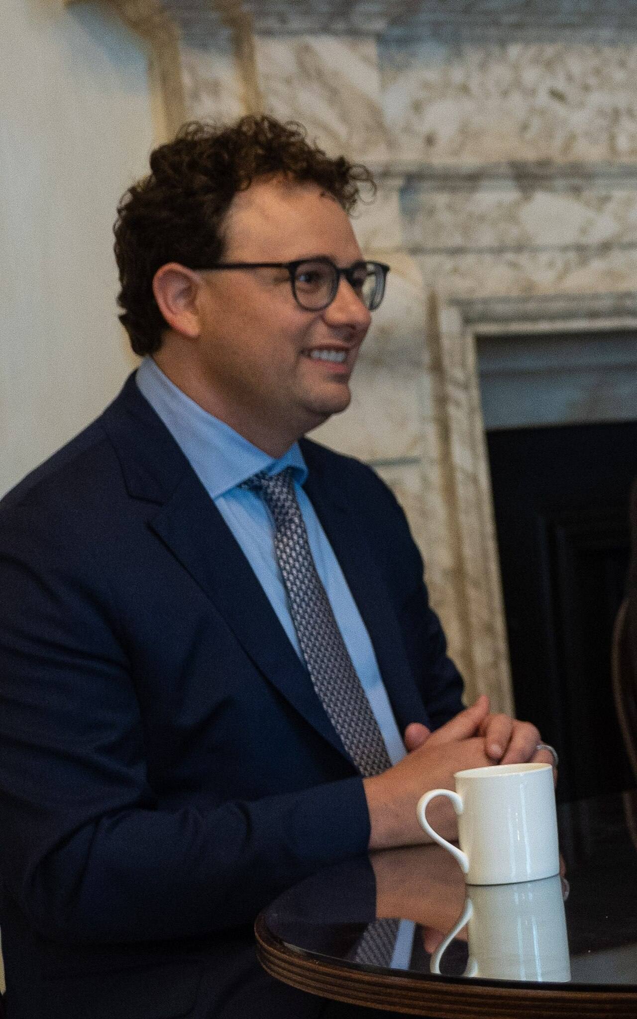 Dario Amodei, consejero delegado de Anthropic, sentado de perfil en un salón del número 10 de Downing Street: traje azul marino, camisa celeste y corbata gris de topos, las manos juntas sobre una mesa redonda con una taza de té blanca, y detrás una chimenea de mármol claro. Fotografiado el 24 de mayo de 2023, durante la reunión del primer ministro británico Rishi Sunak con los responsables de Anthropic, DeepMind y OpenAI.