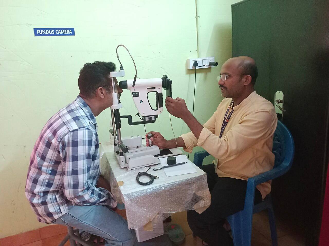 Un técnico sanitario maneja el mando de una cámara de retinografía no midriática mientras un paciente apoya la barbilla y la frente en el soporte del aparato; en la pared, un cartel dice "FUNDUS CAMERA", en una consulta sencilla de India