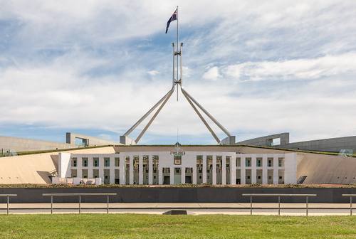 Fachada simétrica del Parlamento de Australia en Canberra, con el mástil y la bandera nacional sobre el tejado, bajo un cielo despejado con nubes.